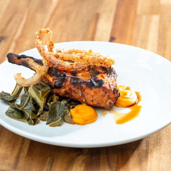 Grilled pork chop with crispy fried onions, collard greens, and a yellow sauce on a white plate.
