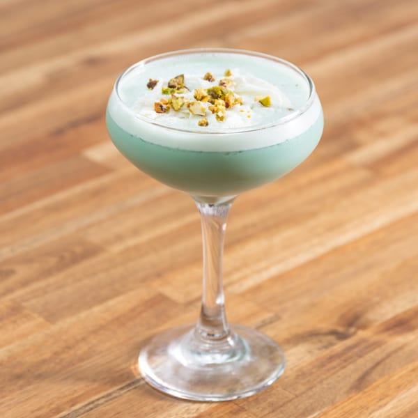 A light green cocktail with white foam and chopped pistachios in a coupe glass on a wooden table.